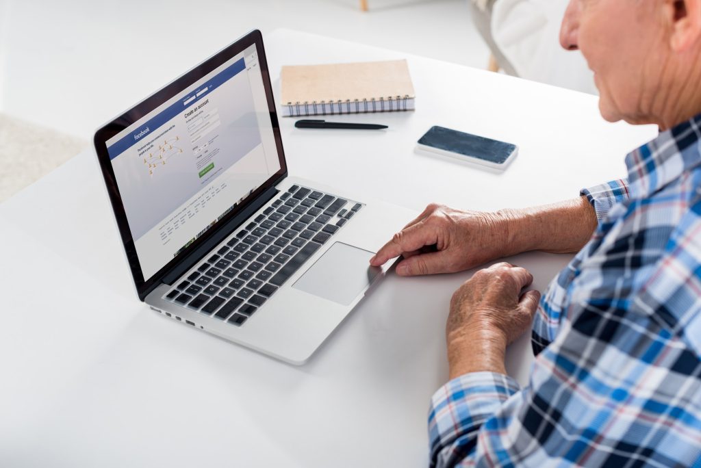 partial view of senior man working on laptop with facebook logo at table with notebook at home